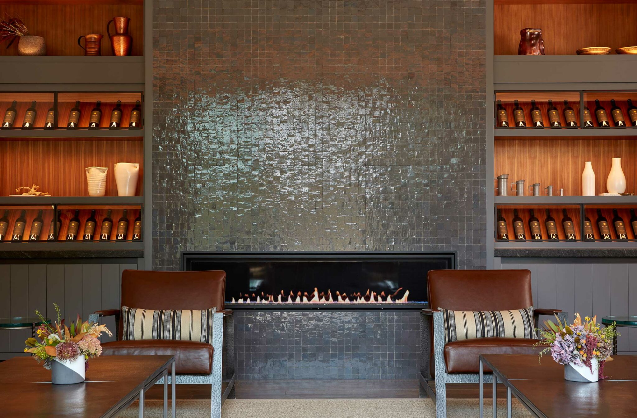 A cozy winery resort lounge with two leather chairs facing a sleek modern fireplace set in a textured dark tile wall. Wooden shelves with vases and bottles flank the fireplace, and floral arrangements add warmth.