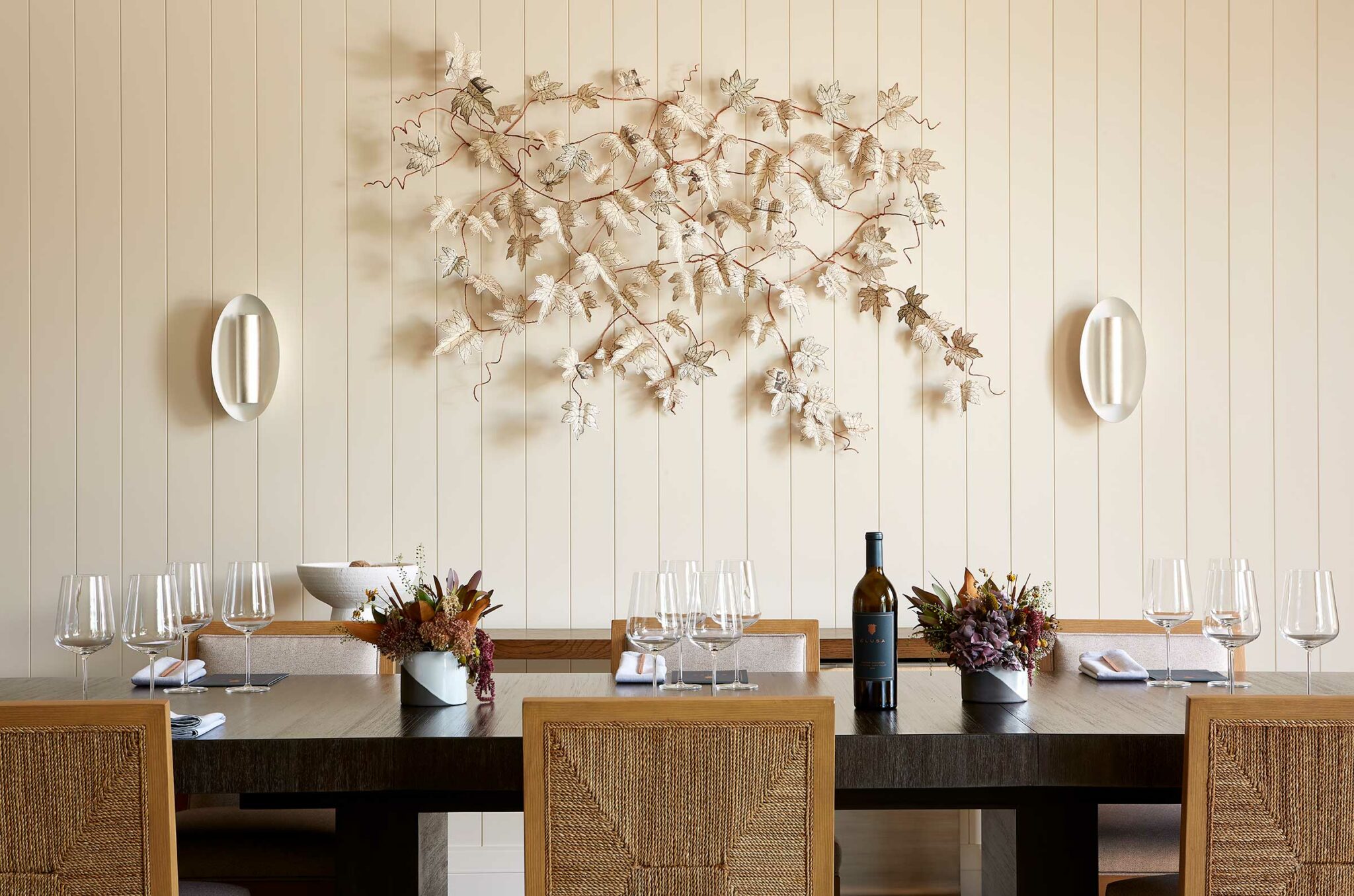 Elegant dining room of a winery resort with a wooden table set for six, featuring wine glasses and a bottle. Wall art of metal leaves adds a natural touch. Warm and inviting.