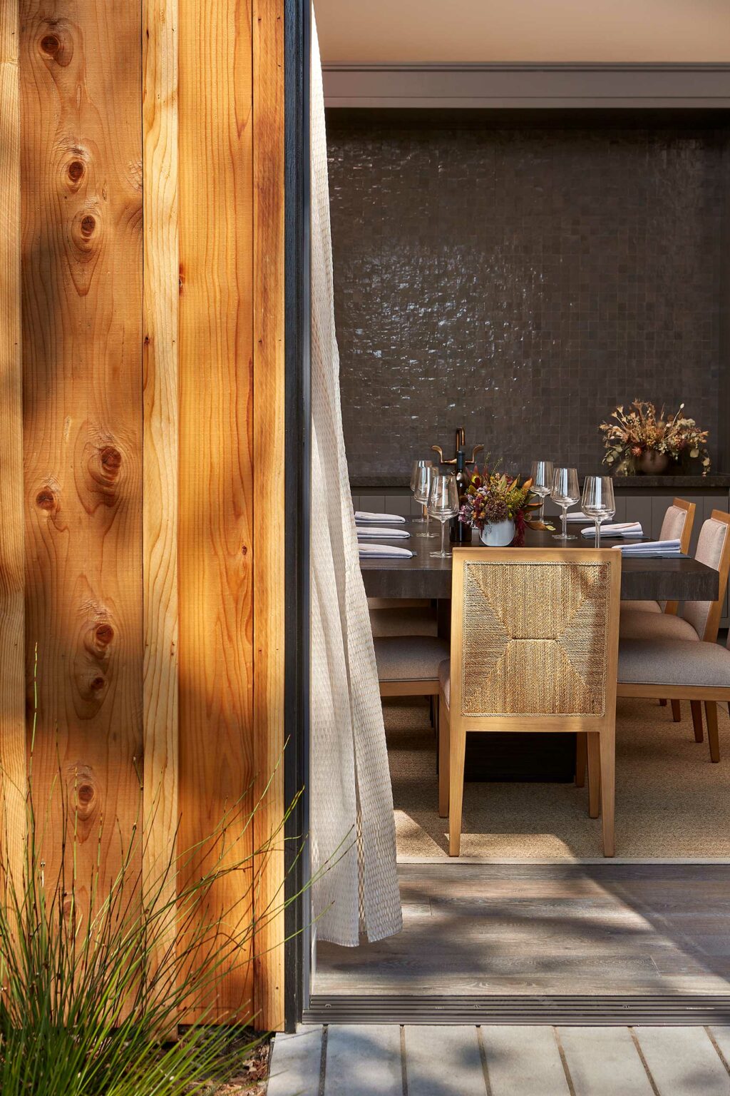 A warm, modern dining room of a winery resort is viewed through a partially open wooden door. The table is elegantly set with glassware and flowers, conveying a cozy, inviting atmosphere.