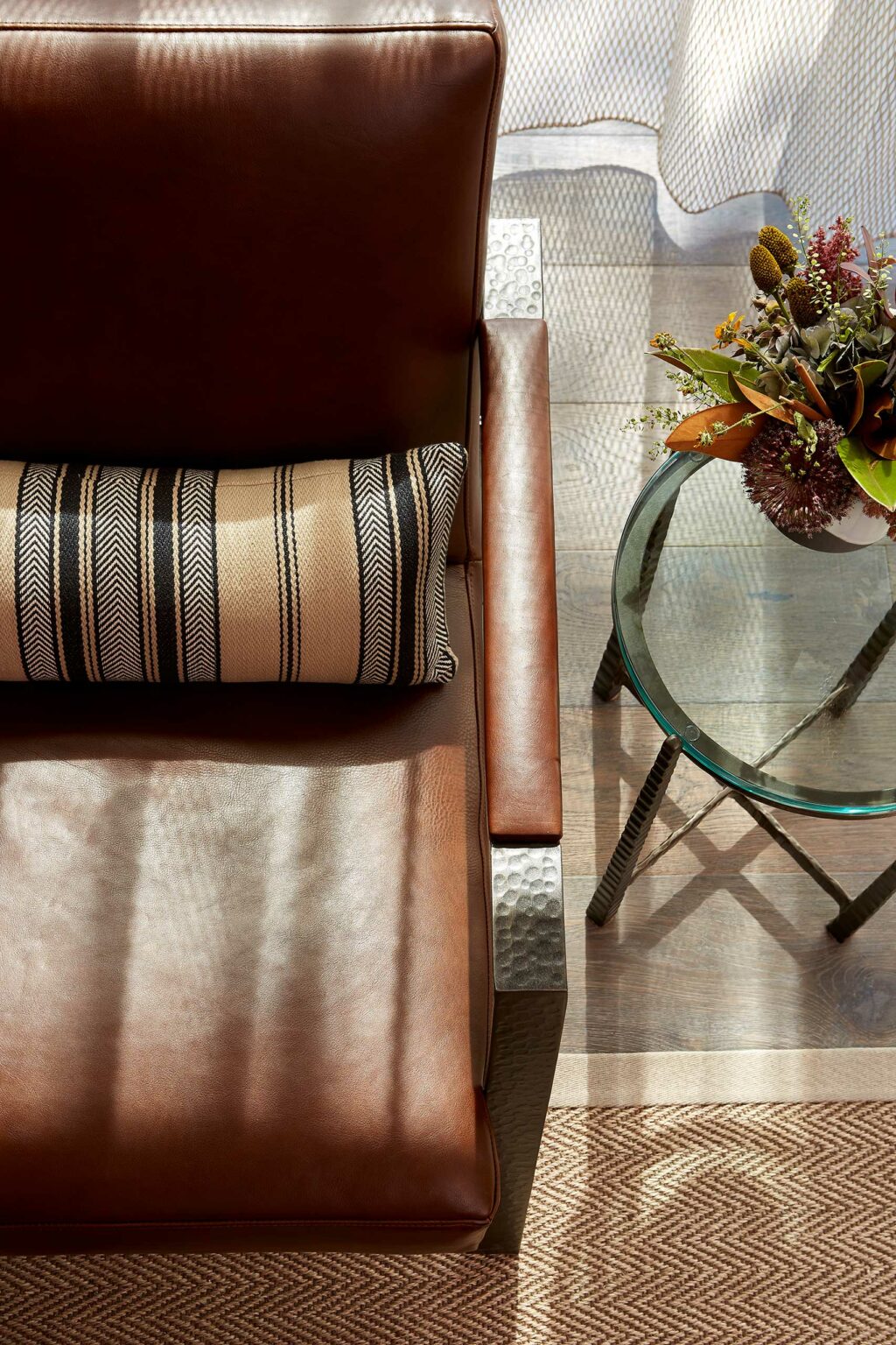 A brown leather chair with a striped cushion beside a glass table topped with a vibrant floral arrangement. Sunlight softly filters across the scene.