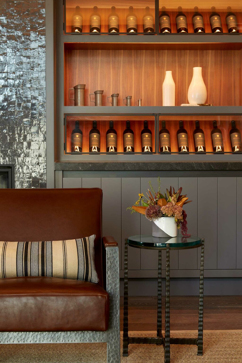 Cozy winery resort room with brown leather chair, striped pillow, and side table holding a floral arrangement. Shelves display bottles, vases, and ceramic cups.