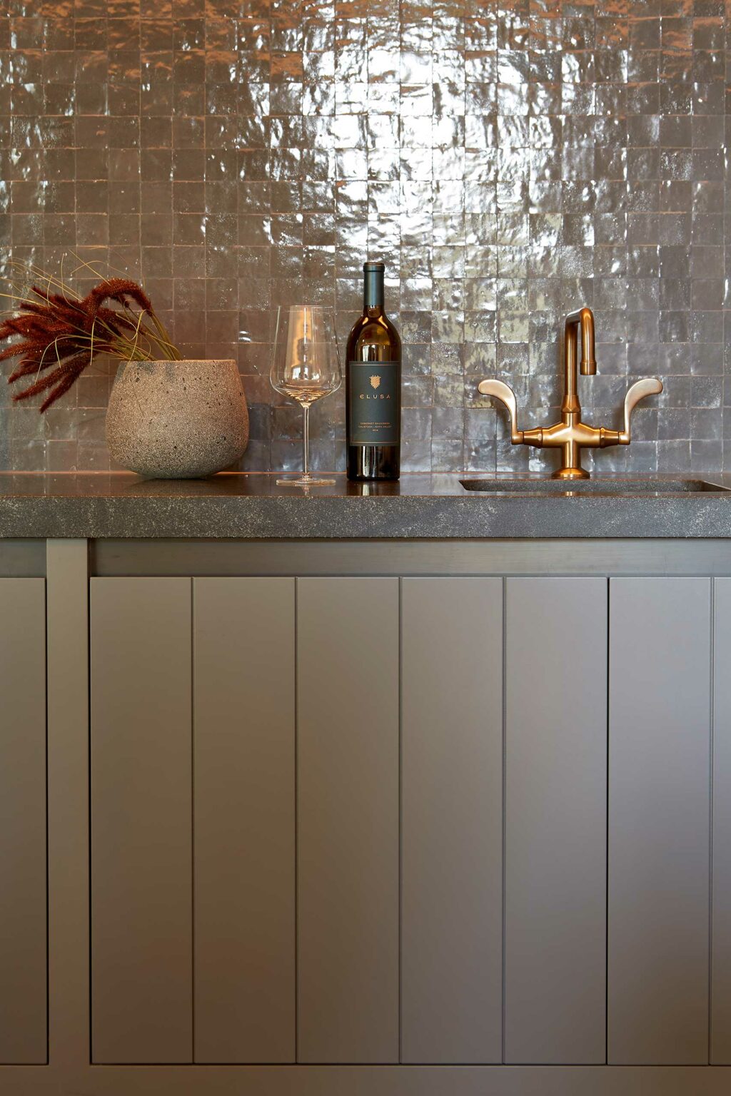 Modern kitchen countertop with a concrete vase holding dried plants, a wine bottle and glass, and a brass faucet against a textured tile backsplash.