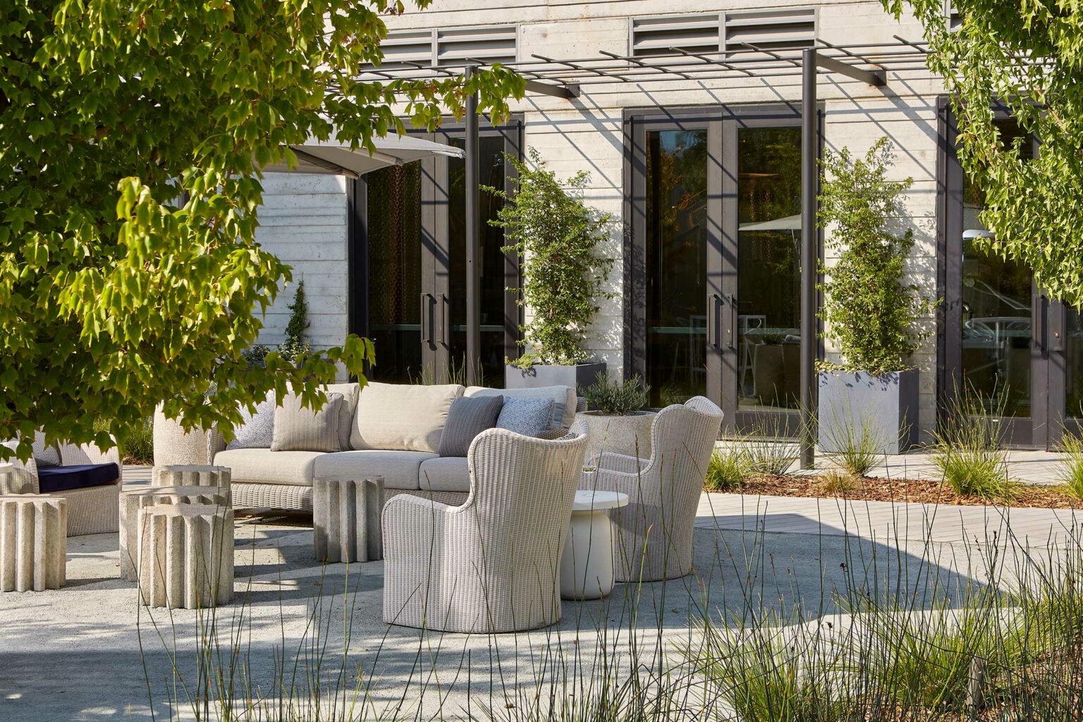 Winery resort outdoor patio with modern woven chairs and a beige sofa around a small table. Surrounded by greenery, it conveys a serene, inviting atmosphere.