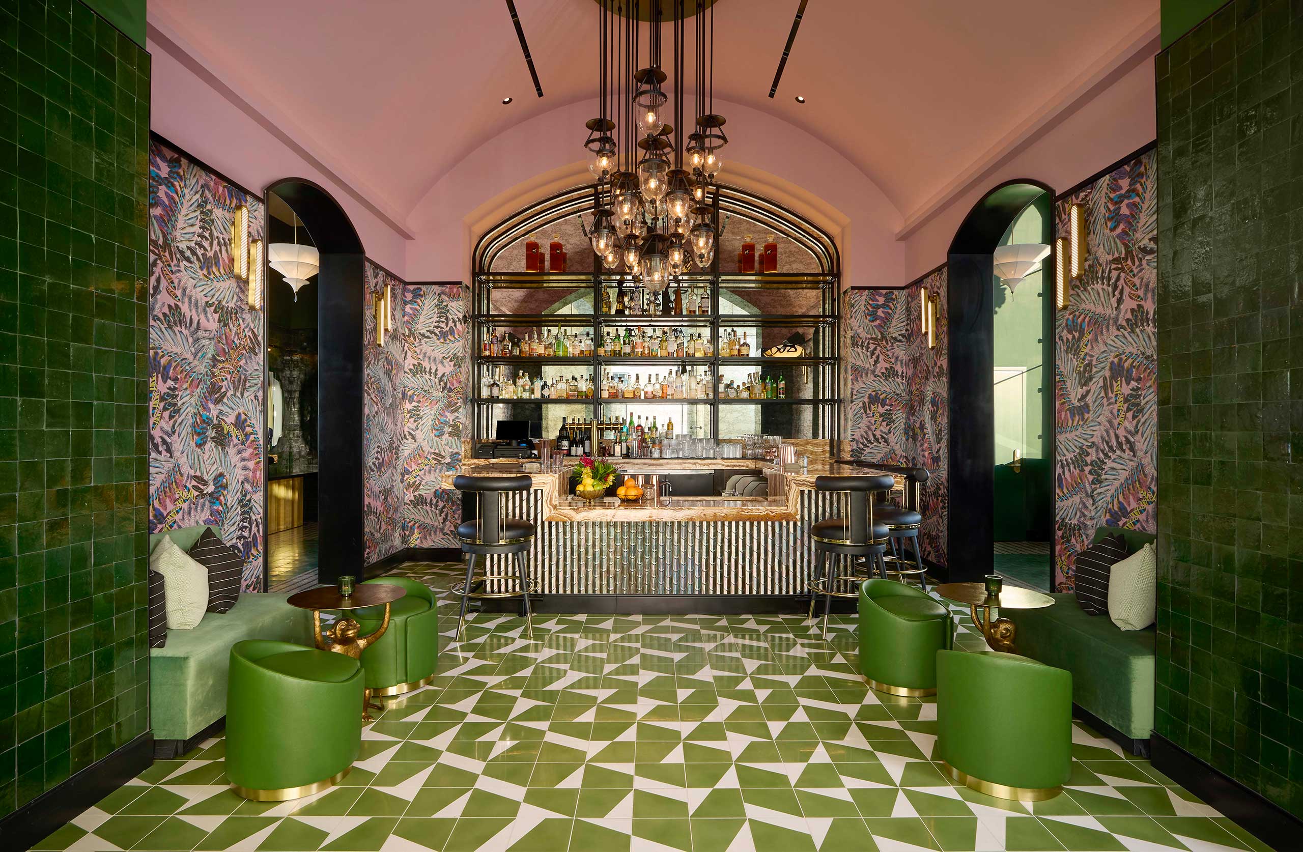 Elegant bar interior of a luxury bar with a mix of green and pink tones. Features patterned wallpaper, geometric green floor, modern chandeliers, and a well-stocked bar.
