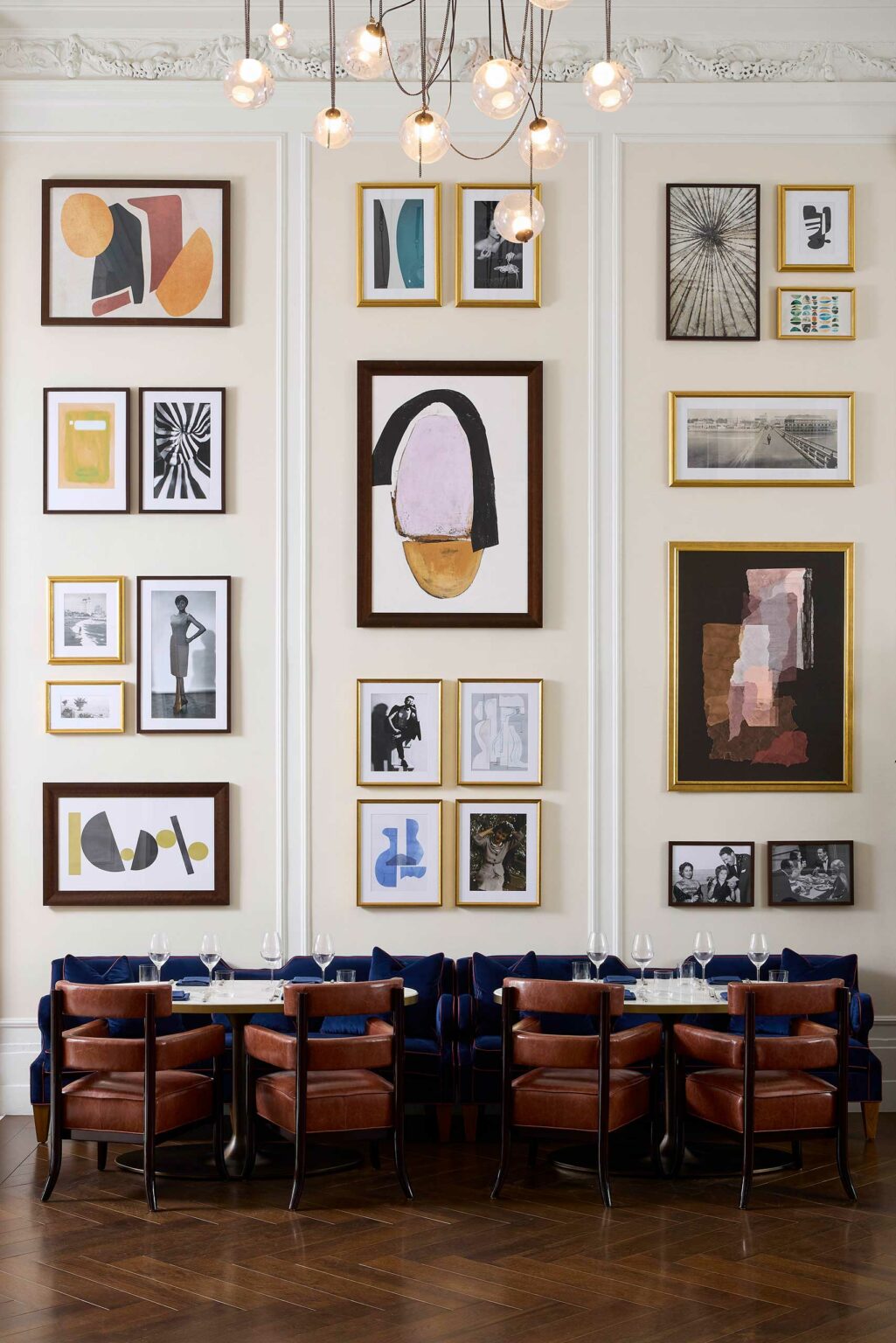 A wall in a chic restaurant of a luxury hotel adorned with an eclectic arrangement of framed abstract and black-and-white art. Below, a table is set with wine glasses.