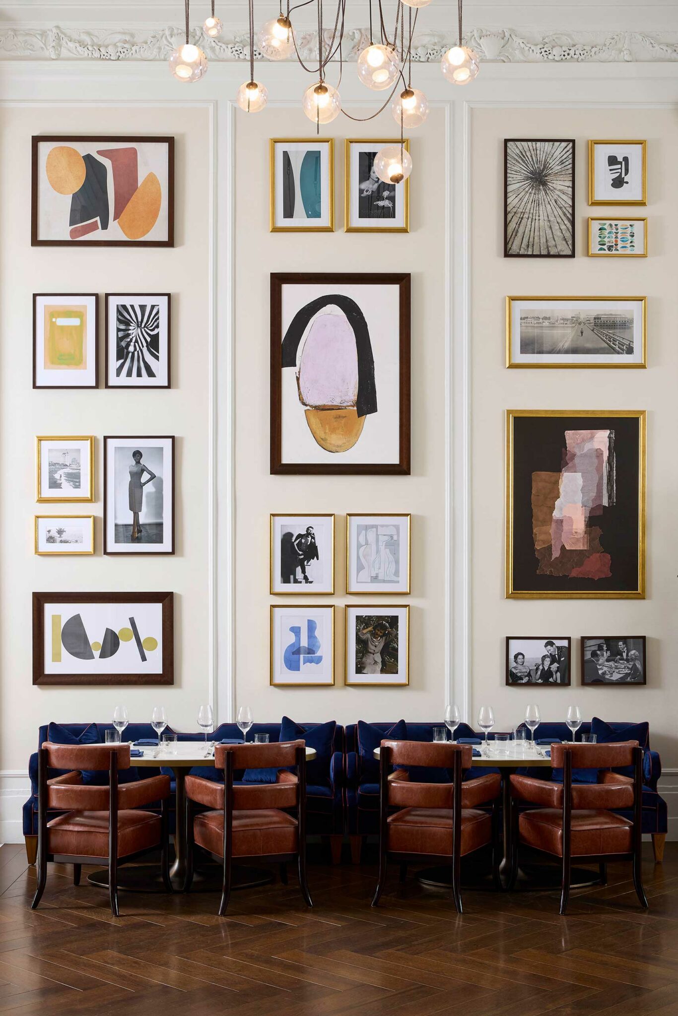 A wall in a chic restaurant of a luxury hotel adorned with an eclectic arrangement of framed abstract and black-and-white art. Below, a table is set with wine glasses.