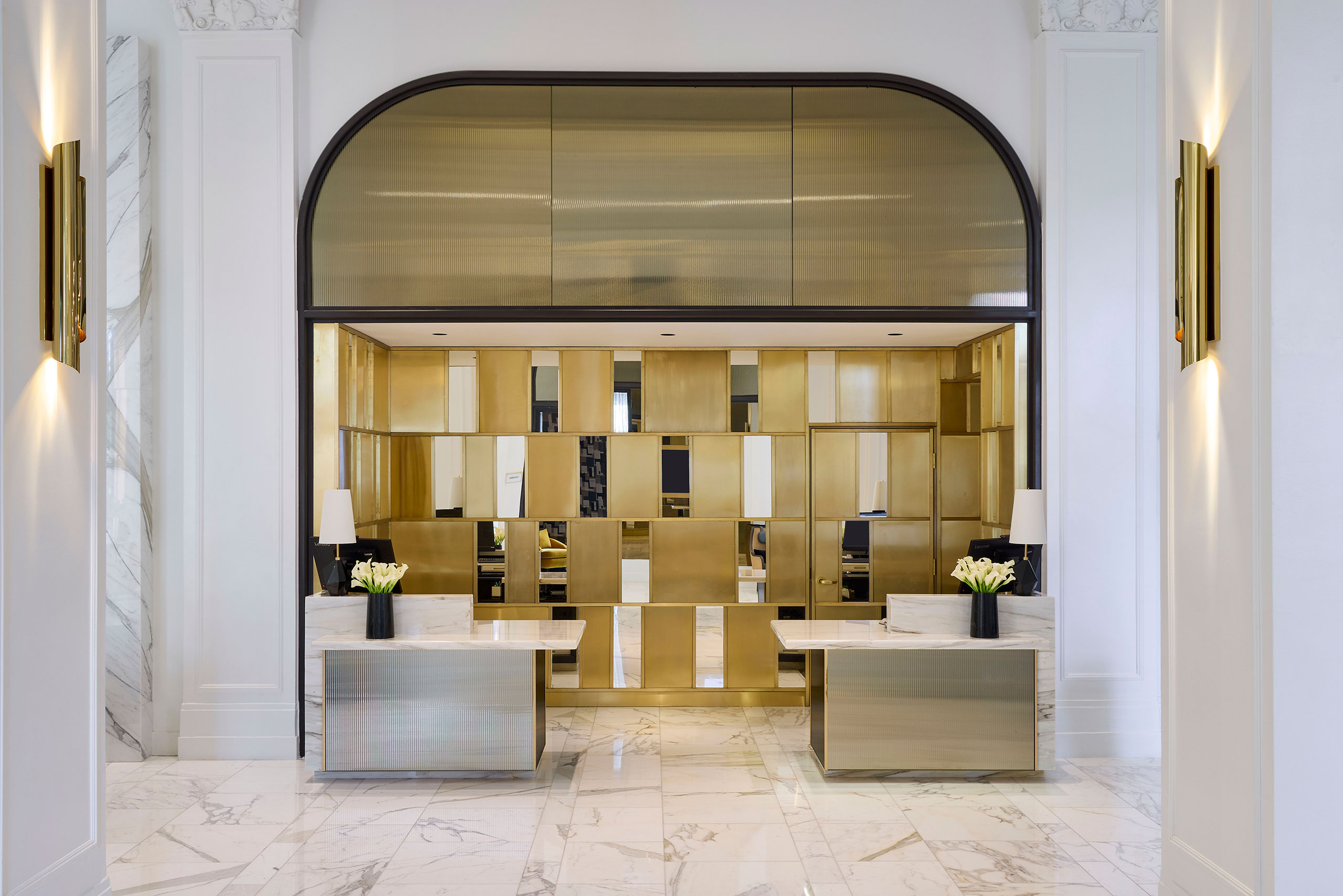 Luxurious hotel lobby with a modern, elegant design. It features two sleek marble desks with gold accents, black vases, and white flowers, creating a sophisticated ambiance.