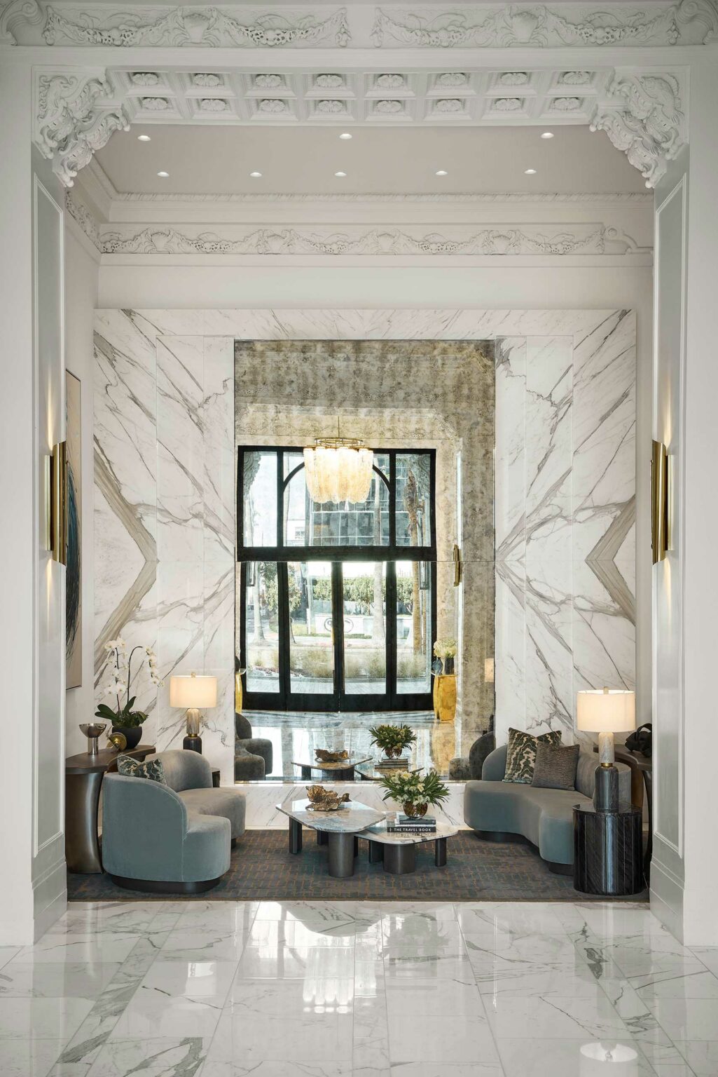 Luxurious hotel lobby with marble walls and intricate ceiling moldings. Features elegant grey seating, lamps, a central glass table, and a large window with a chandelier.