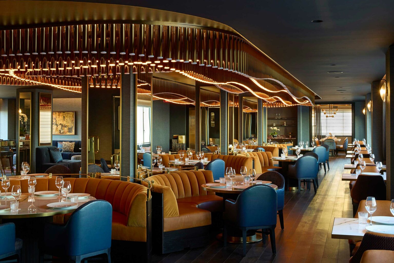 Elegant restaurant interior of a luxury hotel. It features warm lighting, plush orange and blue seating, set tables, and decorative ceiling accents, creating a cozy ambiance.