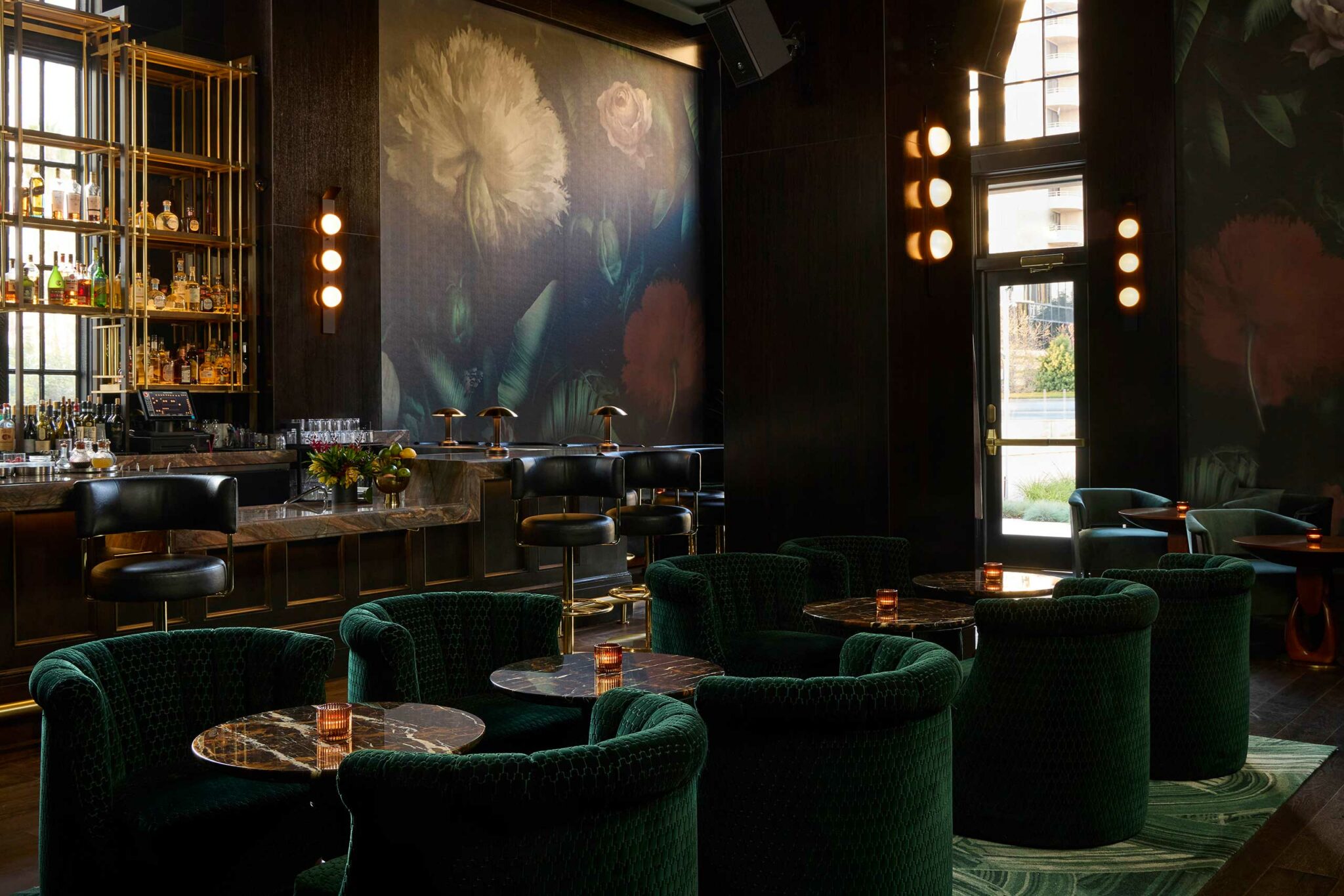 Stylish lounge bar of a luxury hotel with dark floral murals, green velvet chairs, and marble tables. A well-stocked bar and warm lighting create an elegant, cozy atmosphere.