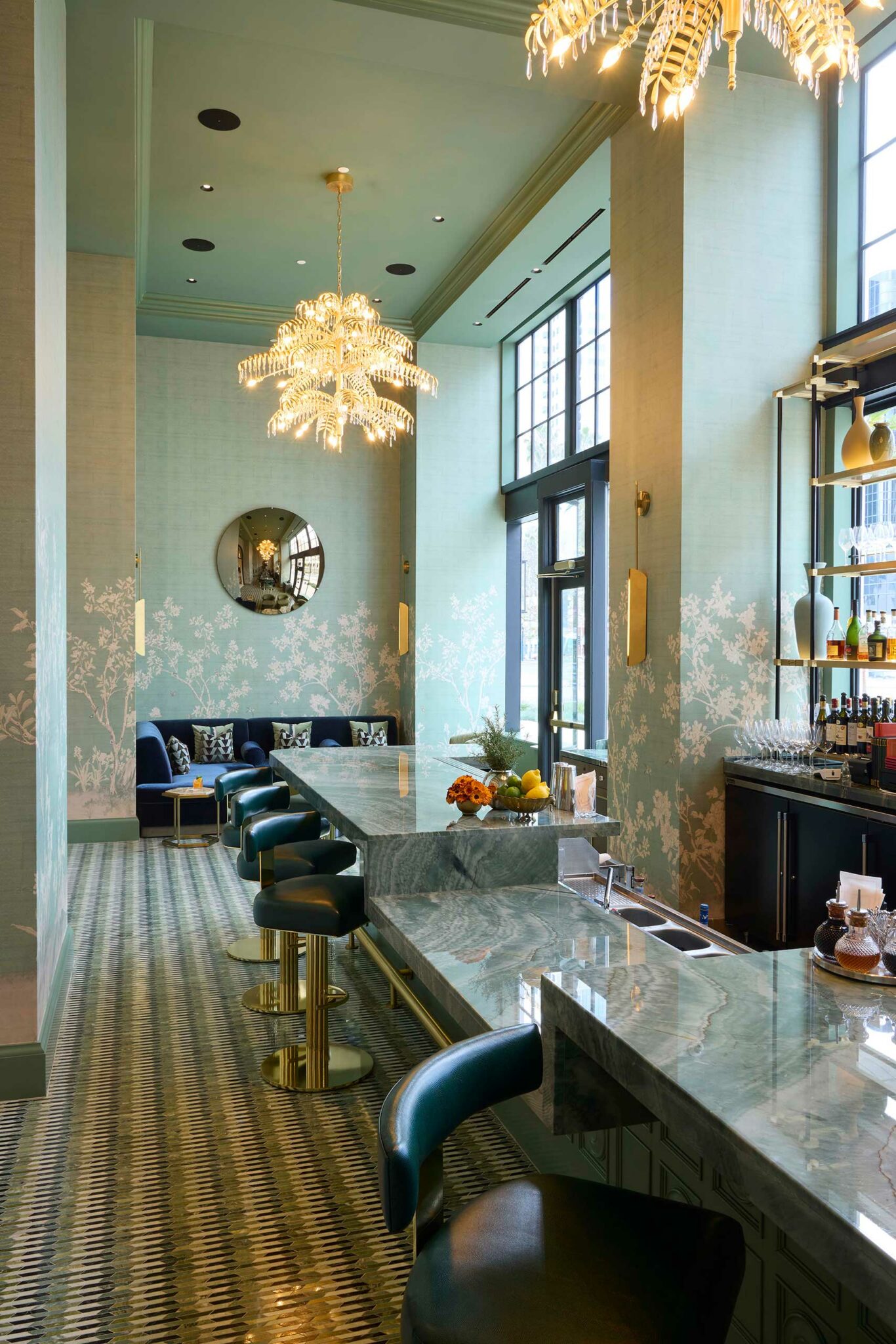 Elegant bar interior of a luxury hotel with teal walls and floral patterns. Marble counters, blue stools, gold accents, and chandeliers create a sophisticated ambiance.