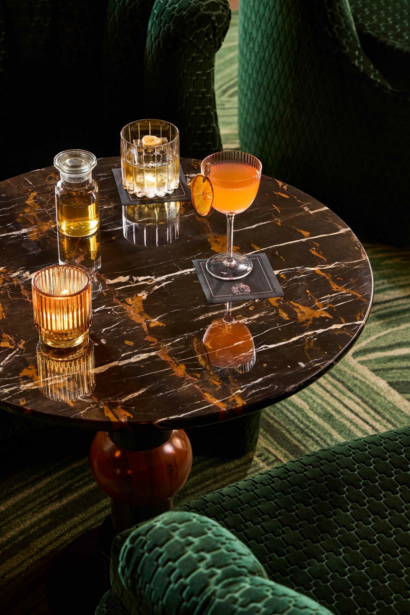 A bar scene in a luxury hotel. It features a marble table with an amber-colored candle, a decanter, a whiskey glass, and a cocktail garnished with an orange slice, surrounded by green chairs.