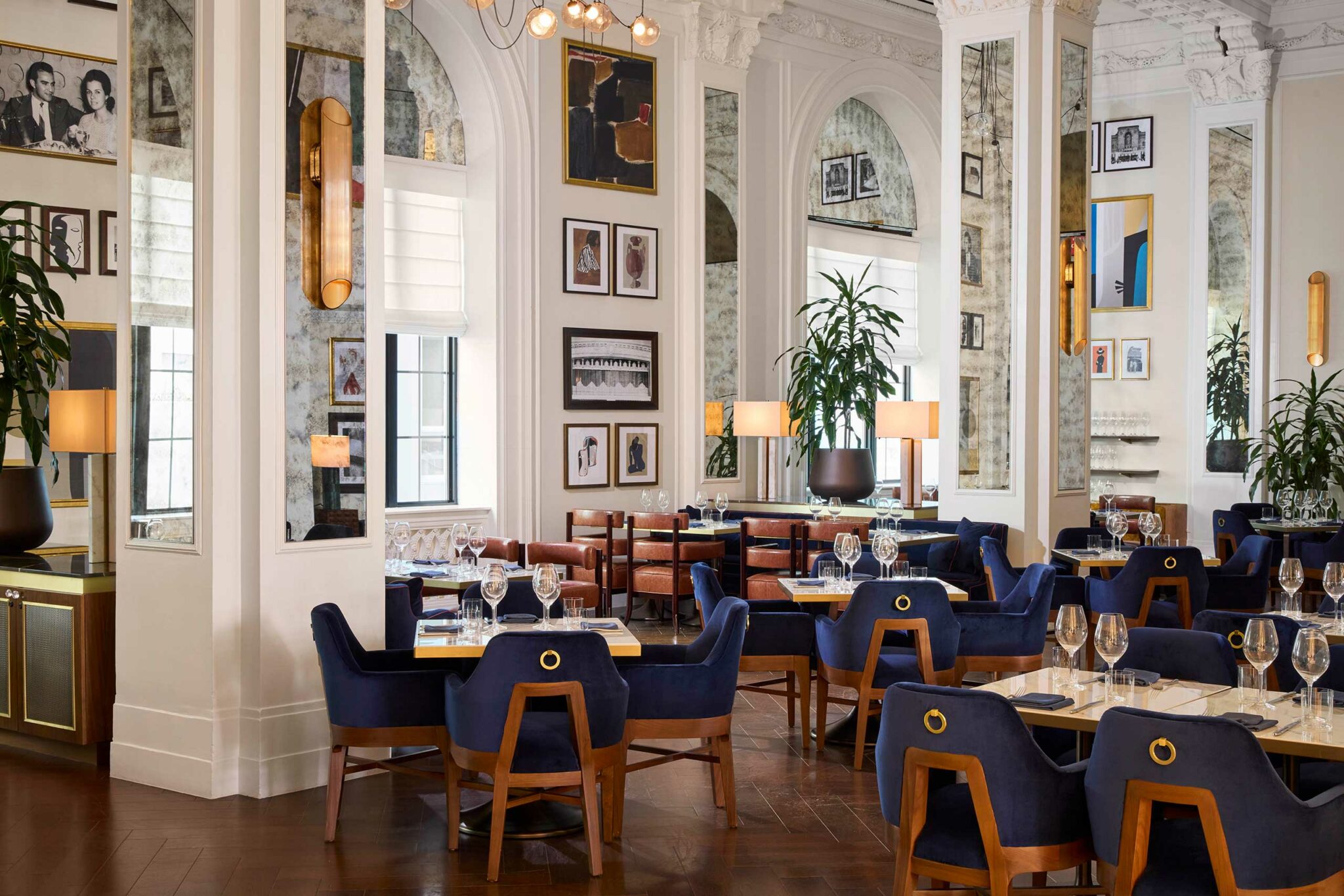 Elegant restaurant interior of a luxury hotel with blue velvet chairs and wood tables set with glasses. Walls adorned with art, mirrors, and lush green plants. Cozy ambiance.