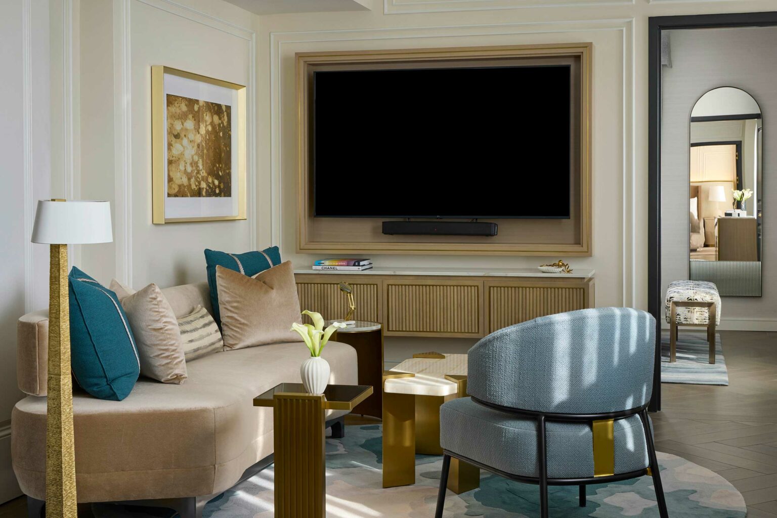 Elegant suite living room with beige sofa, teal pillows, and a modern armchair. A large TV sits in a wooden frame, and a gold-accented table holds flowers.