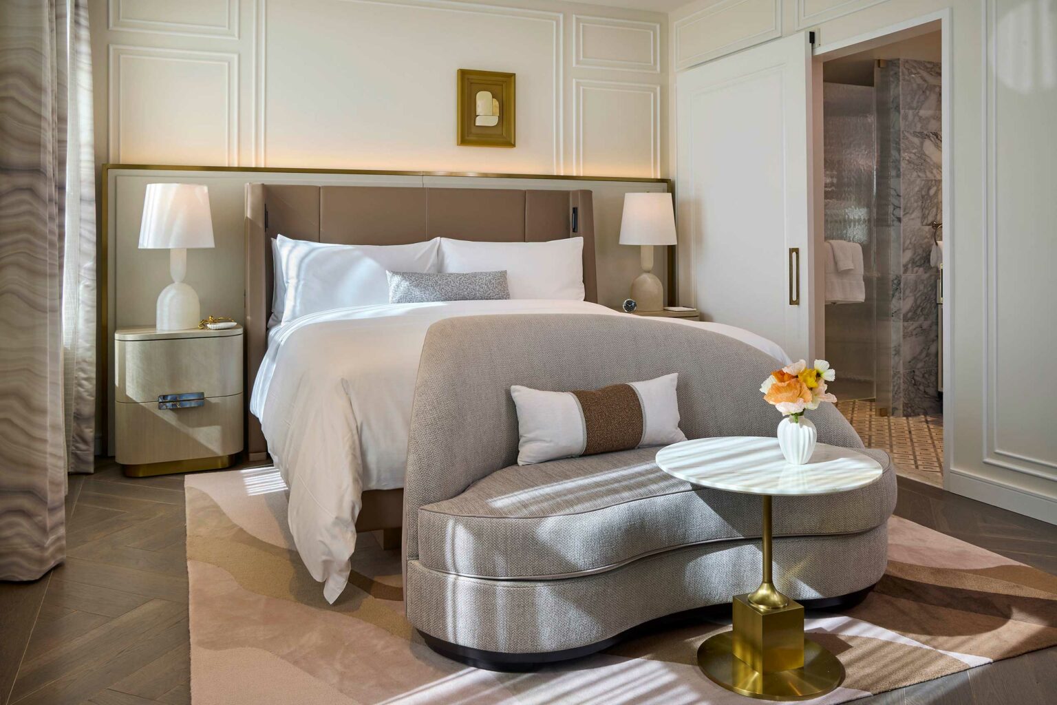 Elegant suite bedroom with a plush bed and crisp white linens, flanked by two lamps on beige nightstands. A cozy gray settee and round table with flowers add charm.
