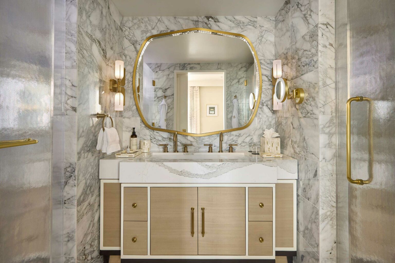 Luxurious suite bathroom with a marble countertop and gold accents. A large, ornate mirror reflects soft light from modern sconces, creating an elegant ambiance.
