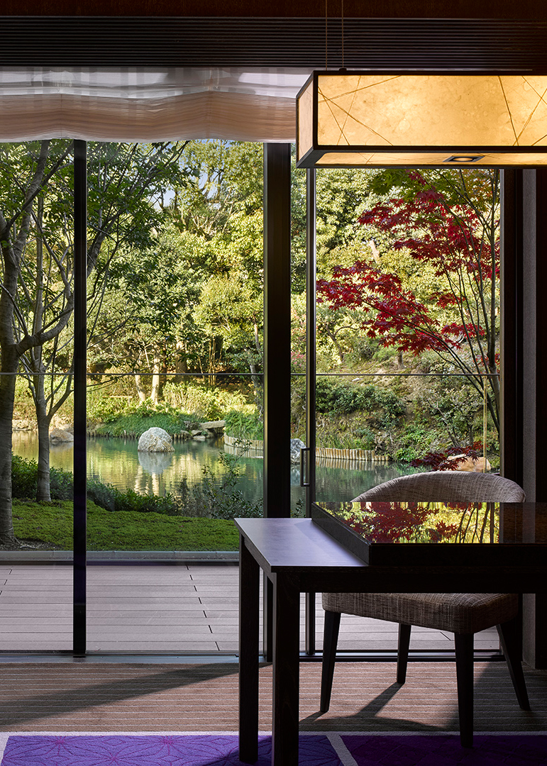 Modern Japanese interior design featuring a dark lacquered wood dining table and chair overlooking a serene Japanese landscaped garden with a pond and autumn foliage.