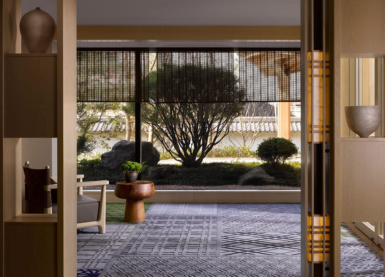 Calm interior hotel space with Japanese garden view. Wooden furniture, patterned rug, and bonsai in foreground; lush greenery and rocks seen through large windows.