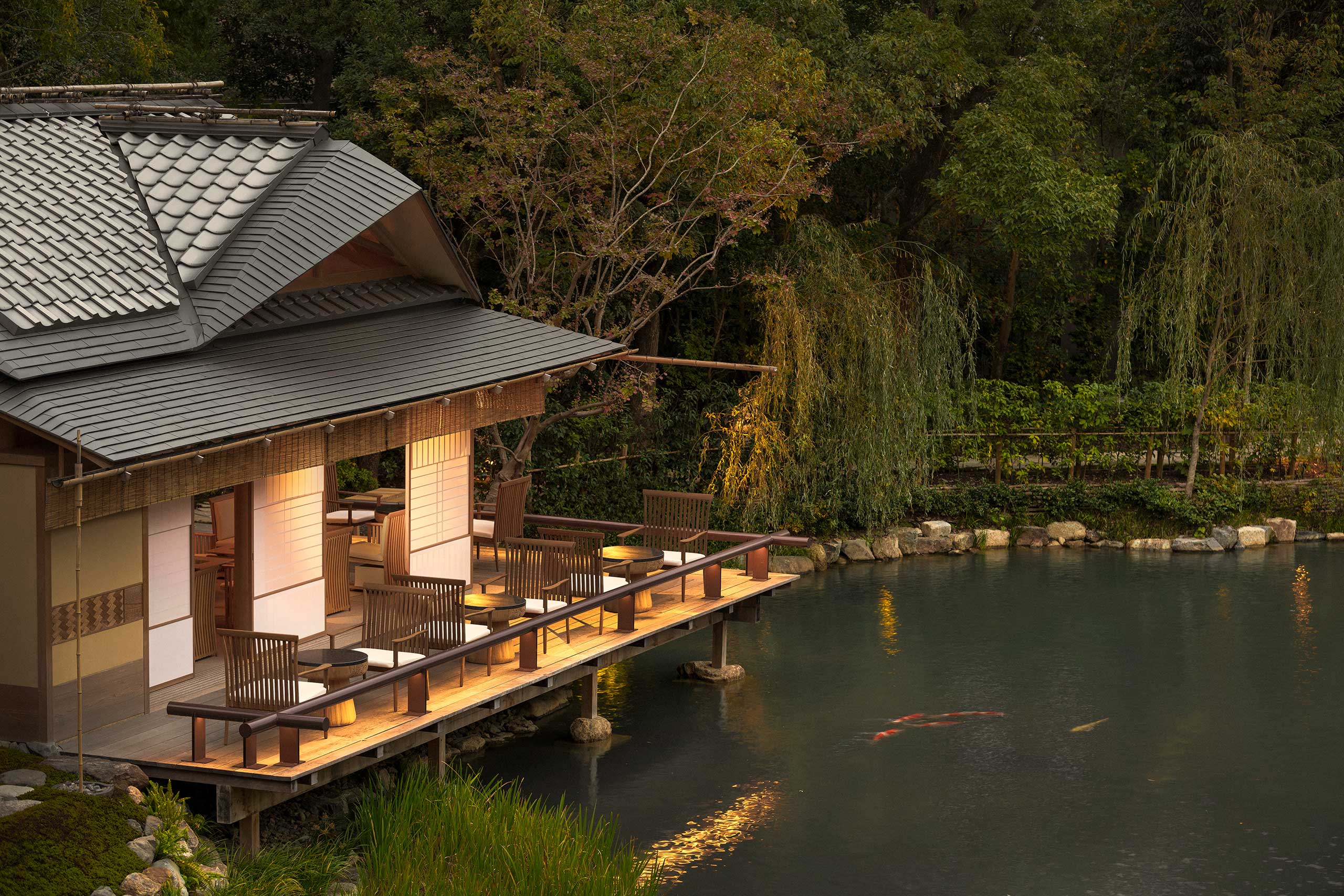 A serene Japanese hotel tea house with a tiled roof and open sliding doors overlooks a tranquil pond. Wooden chairs line the deck, surrounded by lush greenery and koi fish glide in the water.
