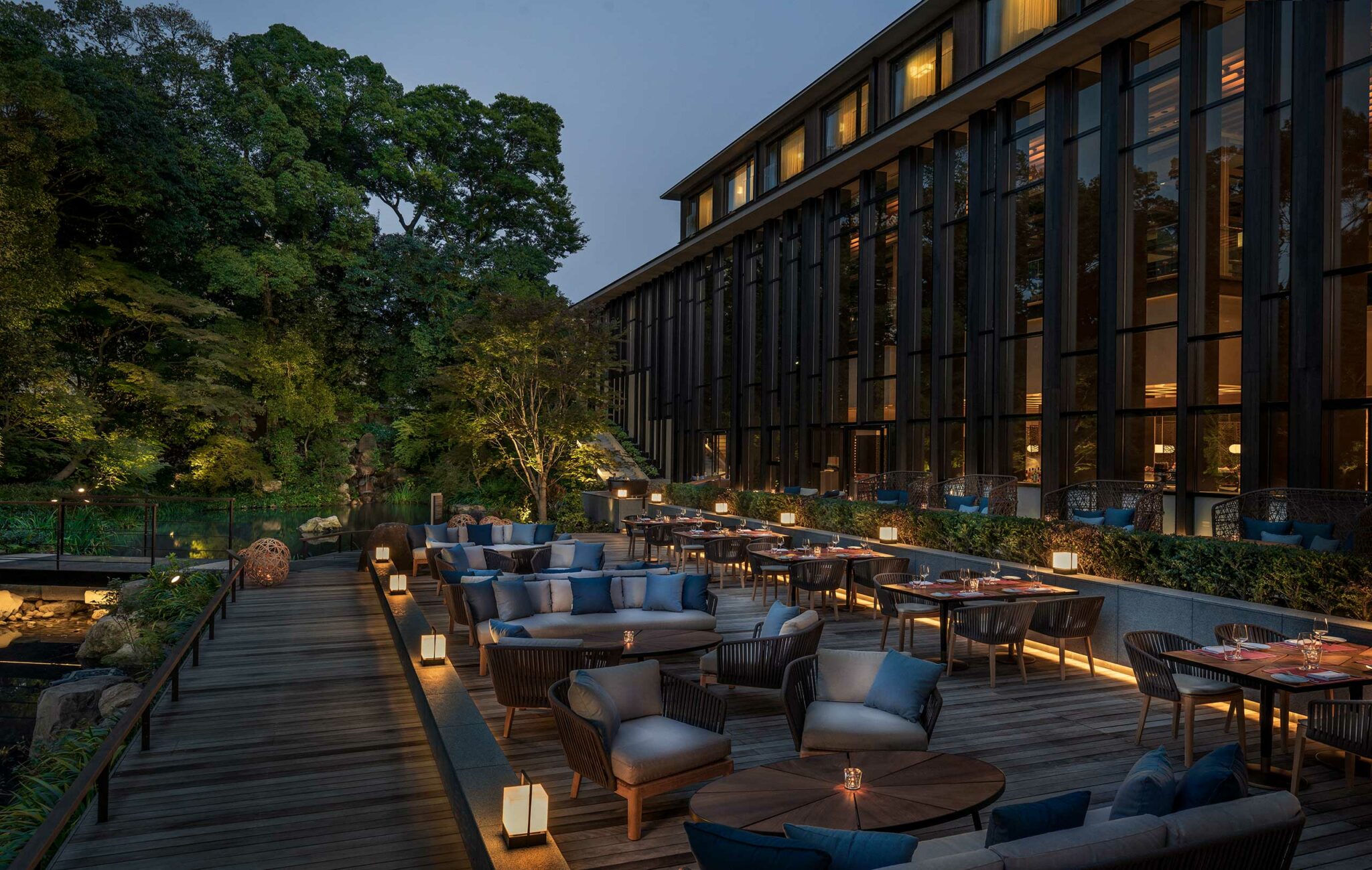 Elegant outdoor hotel restaurant at dusk with cozy seating, candlelit tables, and a serene garden view. Glass building reflects a warm, inviting ambiance.