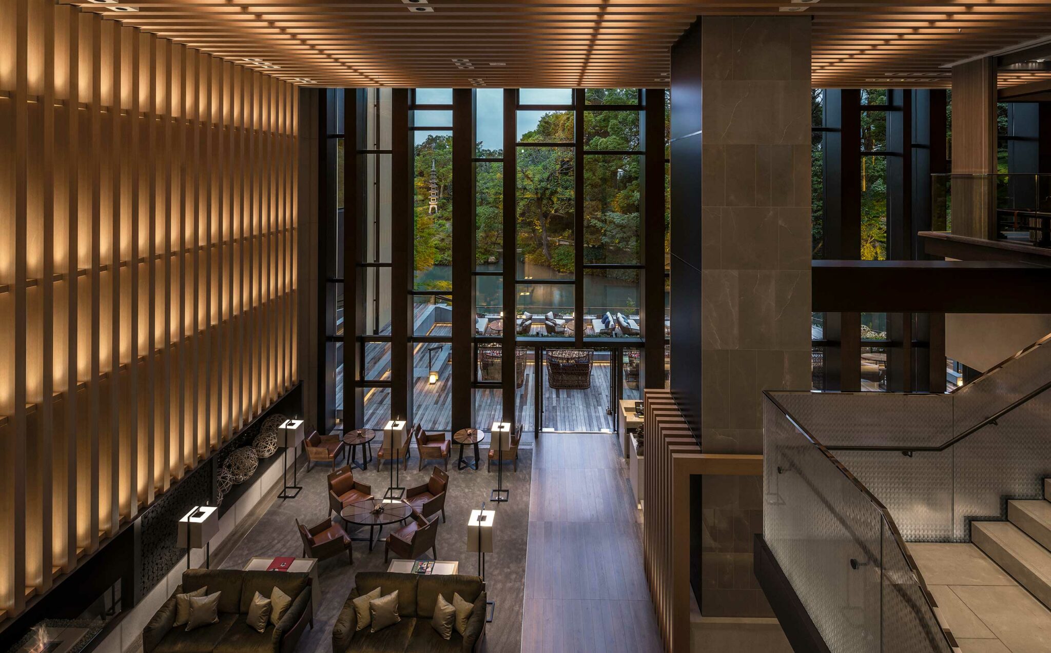 Modern hotel lobby with tall glass windows, warm lighting, and elegant wooden paneling. Cozy seating areas and lush greenery visible outside. Calm atmosphere.