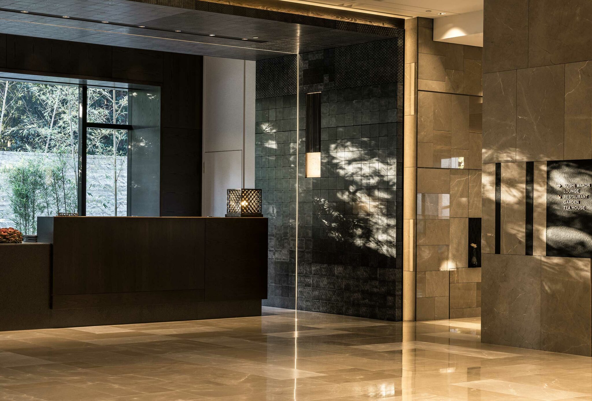 Elegant hotel lobby with a dark, sleek reception desk, modern lighting, reflective stone walls, and dappled sunlight creating a serene atmosphere.