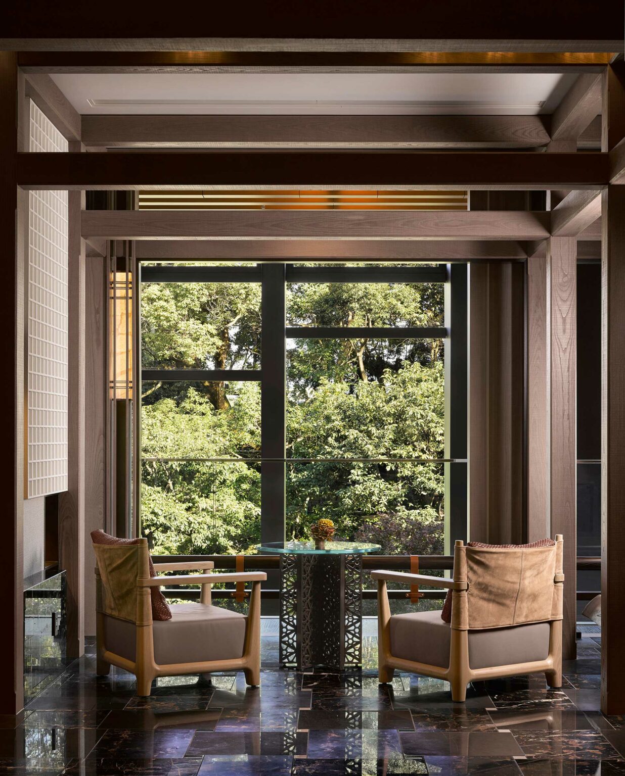 Two wooden armchairs face a large window overlooking lush green trees. A small glass table sits between them, creating a tranquil, inviting atmosphere within a hotel setting.