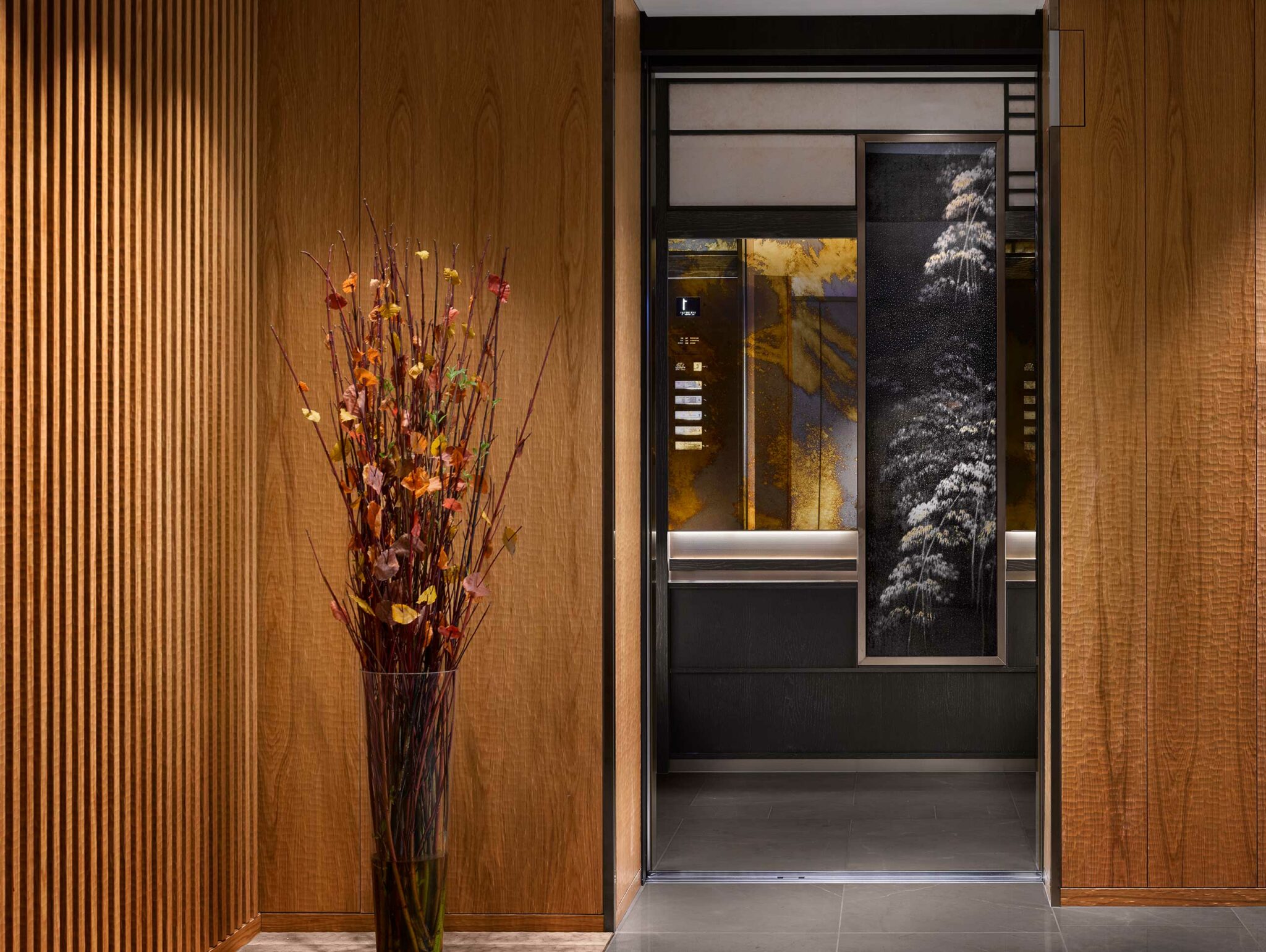 A modern hotel elevator with wood paneling, intricate floral artwork, and warm ambient lighting. A vase with colorful autumn branches stands nearby.