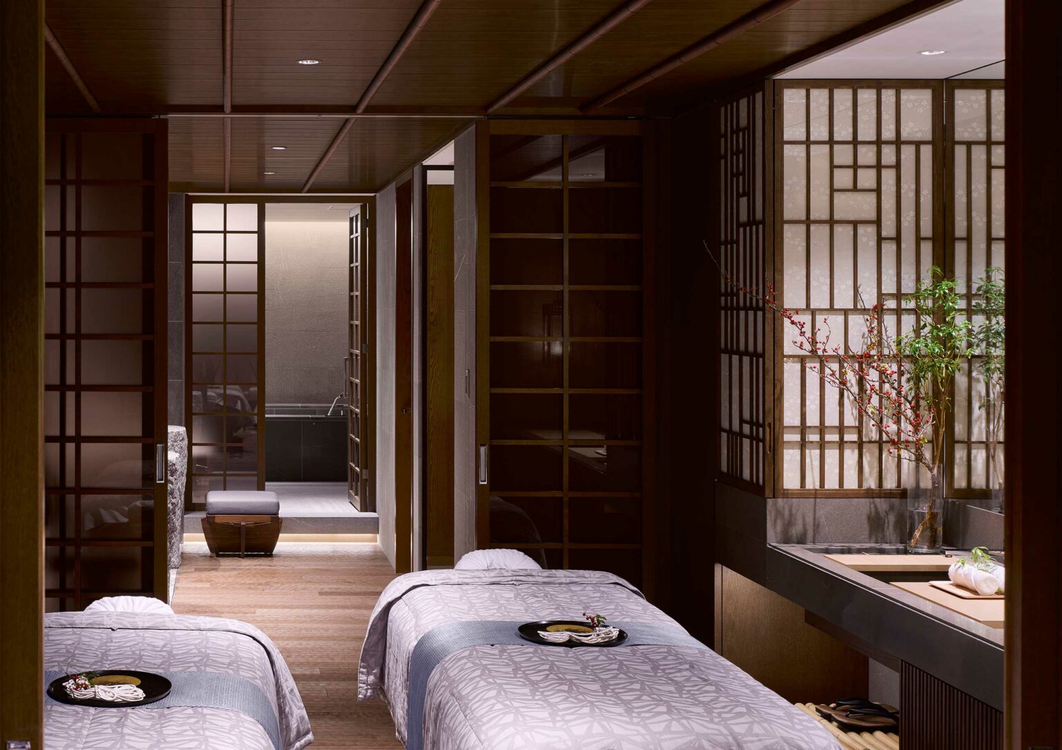 Tranquil hotel spa treatment room with two massage beds, soft lighting, and Japanese shoji screens. A vase with flowers adds elegance, creating a serene ambiance.