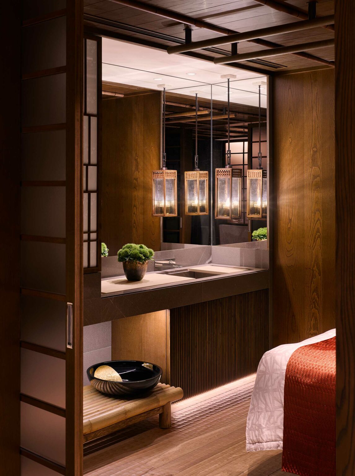 Warm, elegant hotel spa bathroom with wood accents. Soft lighting from hanging lanterns reflects in a mirror. A potted plant and black bowl add decor. Calm ambiance.