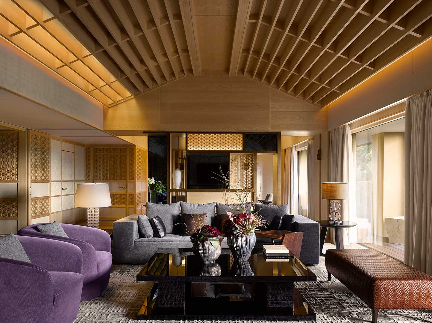 Elegant hotel lounge with purple armchairs, gray sofa, and decorative plants on a glossy coffee table. Warm lighting and textured walls create a cozy ambiance.