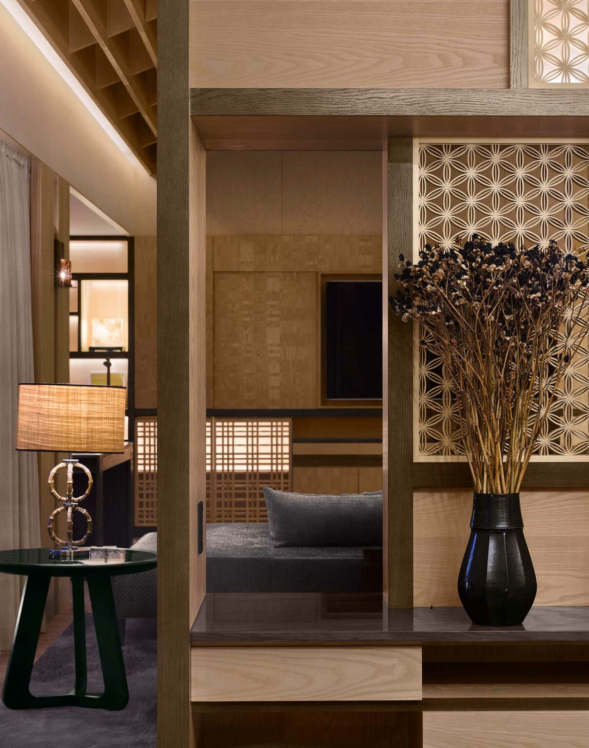 Modern suite living room with wooden accents and soft lighting. A decorative lamp and dried flowers in a vase sit on a table, exuding a cozy, serene atmosphere.