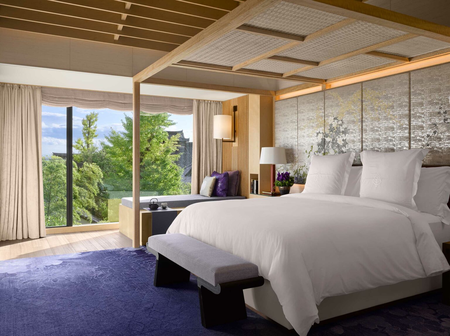 Luxurious hotel bedroom with white bedding, wooden canopy, and purple accents. Large window reveals lush greenery. Serene, elegant atmosphere.