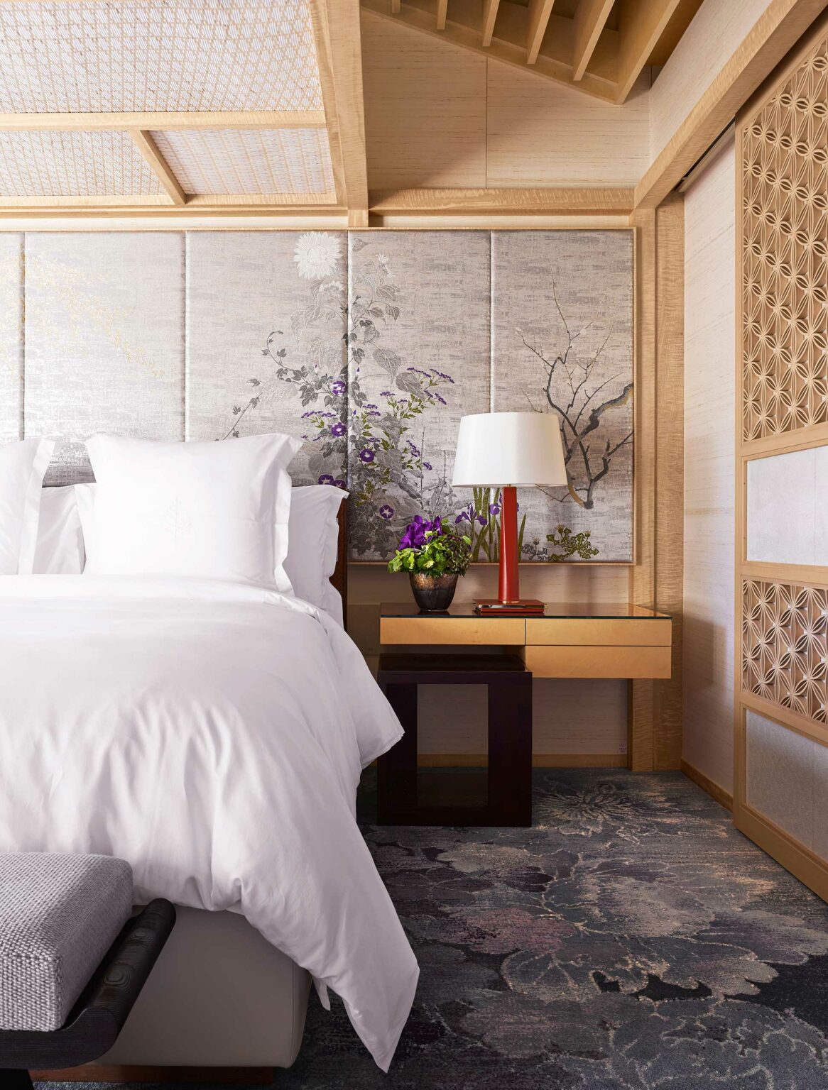 Elegant hotel bedroom with a white bed, wooden accents, and a floral Asian-style mural. A red lamp and purple flowers sit on a light wood nightstand, conveying serenity.