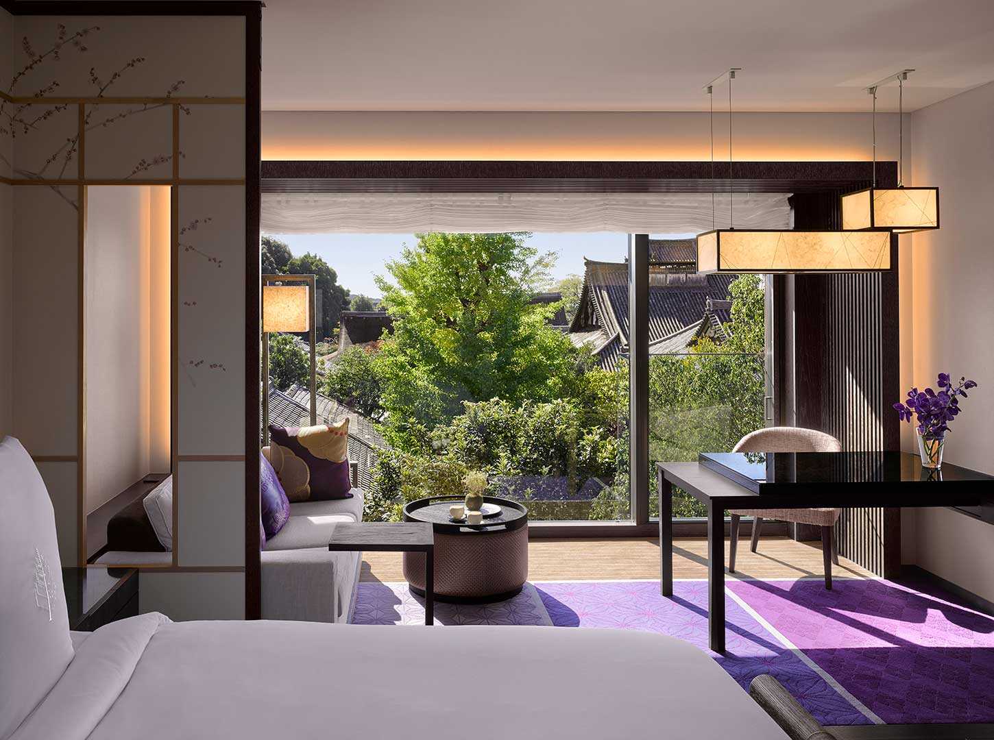 Elegant hotel room with large window view of lush greenery and traditional rooftops. Modern decor features purple accents and a cozy ambiance.