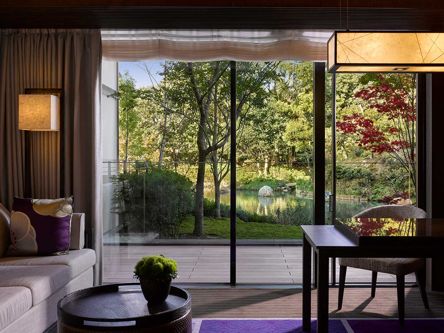 Modern suite living room with large glass doors opening to a serene garden with trees and a pond. Soft lighting and a cozy ambiance enhance tranquility.
