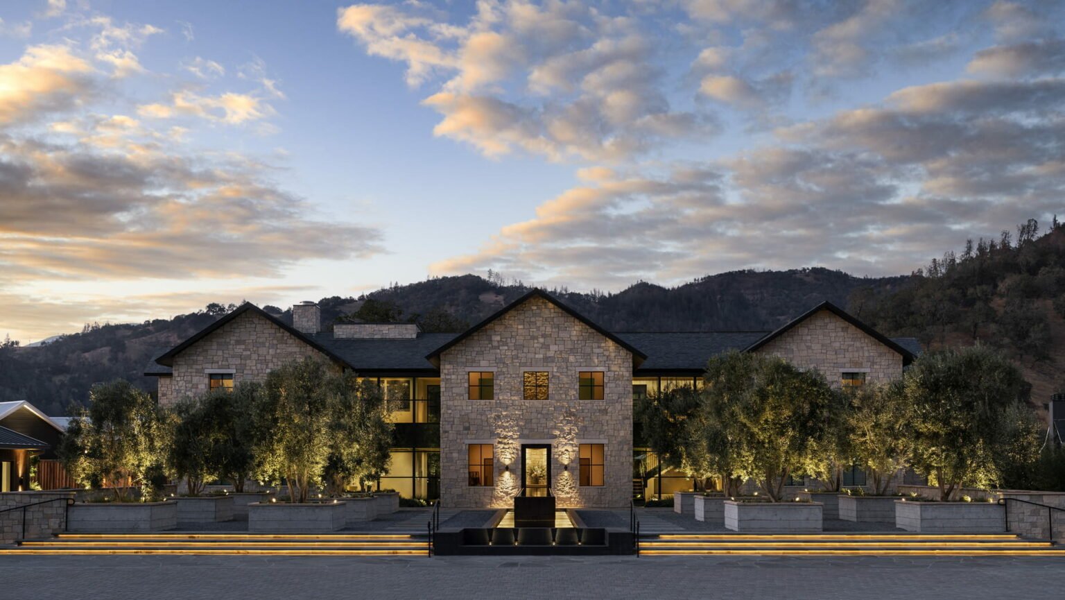 Exterior of Four Seasons Resort Napa Valley. It features a two-story stone building at dusk with soft lighting and symmetrical design, surrounded by trees. Mountains and a partly cloudy sky in the background. Luxurious ambiance.