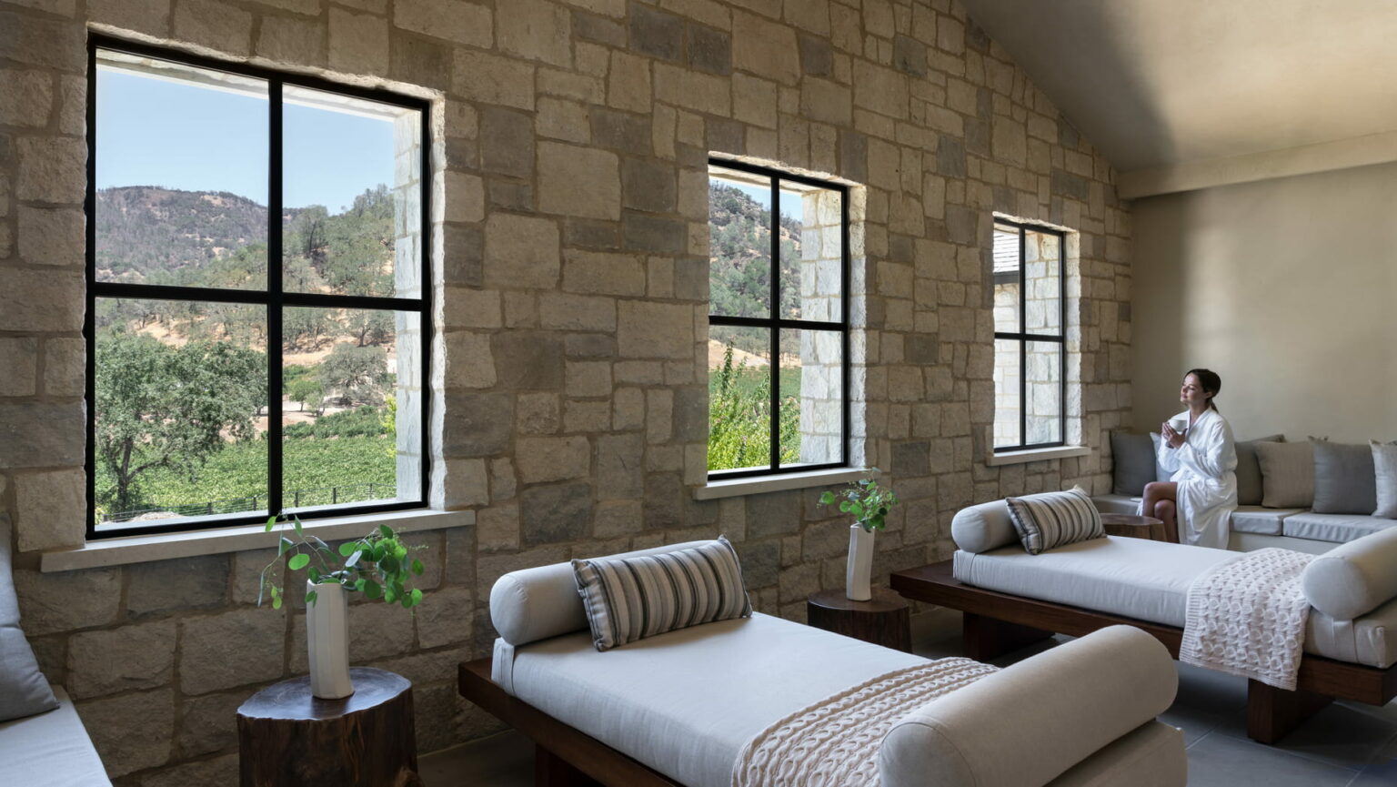 Luxurious resort spa room with stone walls and large windows offering a scenic view of mountains and greenery. A person in a robe relaxes on a cushioned lounger.