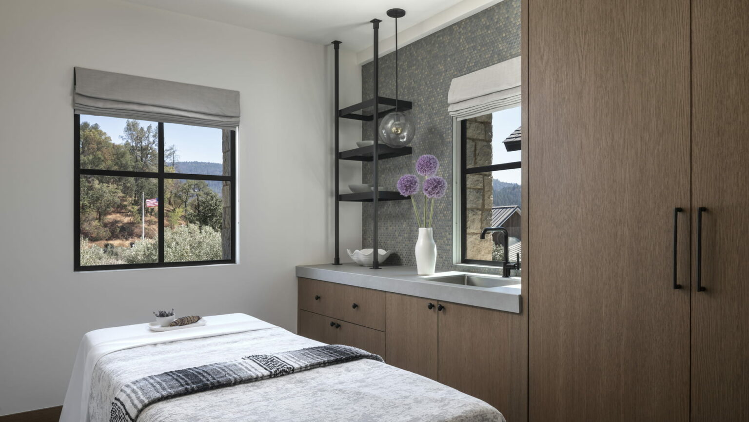 A serene resort spa treatment room with a massage bed and folded towels, adjacent to a countertop with purple flowers. A window shows greenery and hills outside.