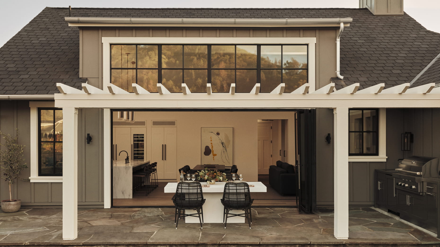 Modern patio with pergola, leading to an open-plan kitchen and living room through large glass doors. Outdoor table with chairs, cozy ambiance.