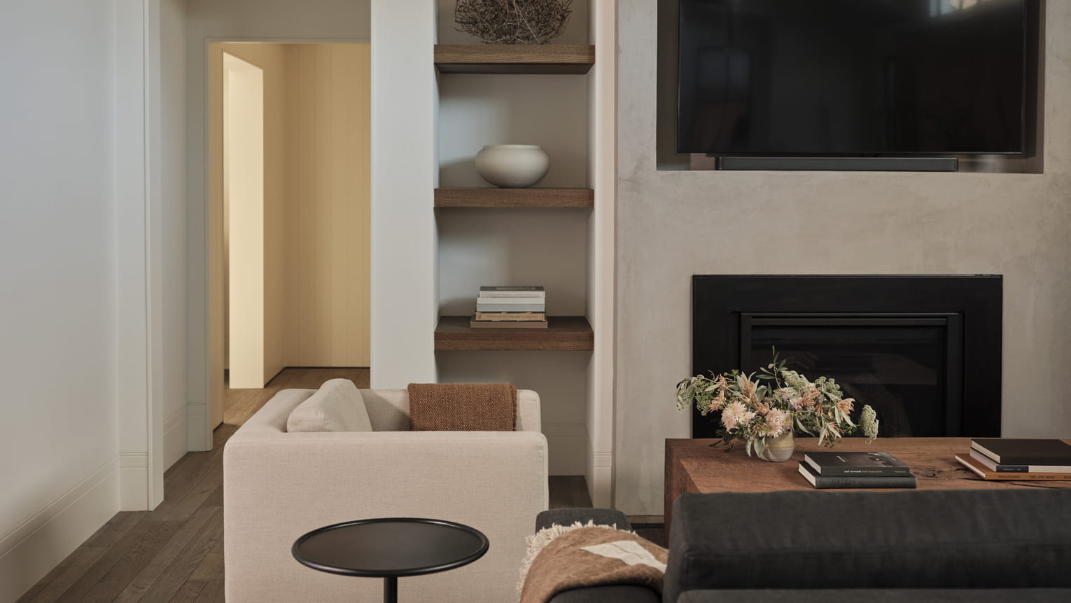 Modern suite living room with neutral tones. Features a beige couch, wooden shelves with decor, a TV above a fireplace, and a table with flowers and books. Cozy and minimalistic feel.