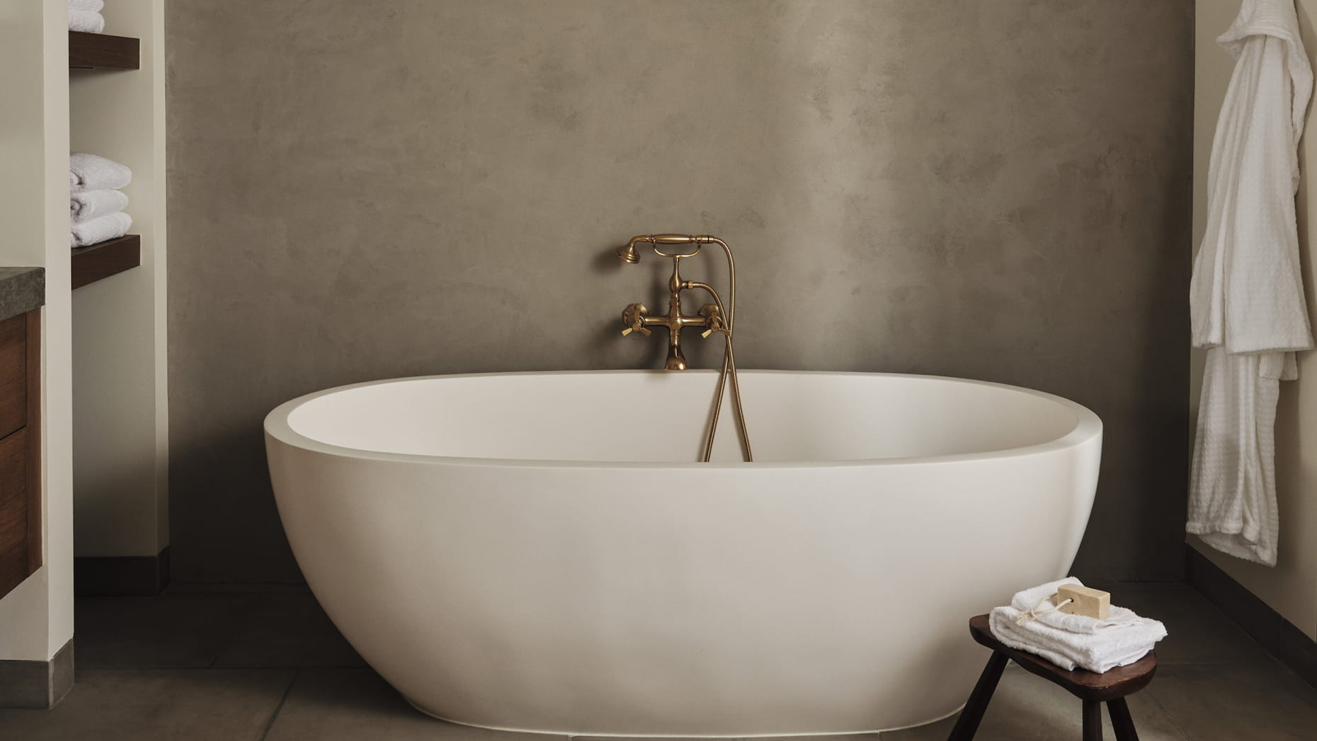 A modern suite bathroom with a freestanding white oval bathtub against a taupe wall, featuring brass fixtures. A small stool holds folded towels, conveying a serene and minimalist ambiance.
