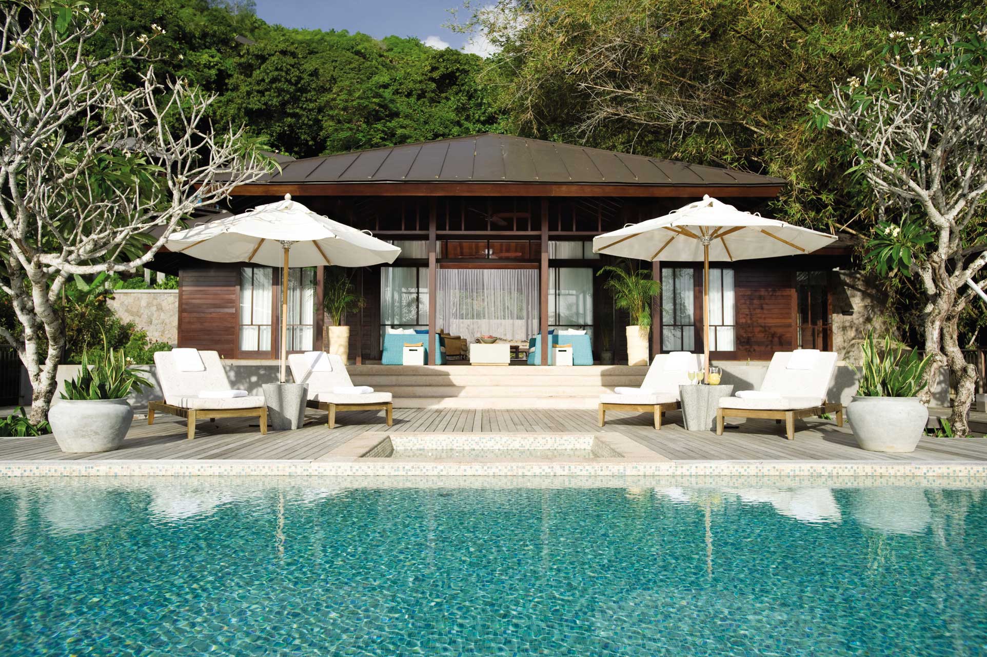 Luxurious poolside scene with a wooden hotel bungalow, surrounded by lush greenery. Four sun loungers and white umbrellas line the pool's edge, creating a serene, inviting atmosphere.