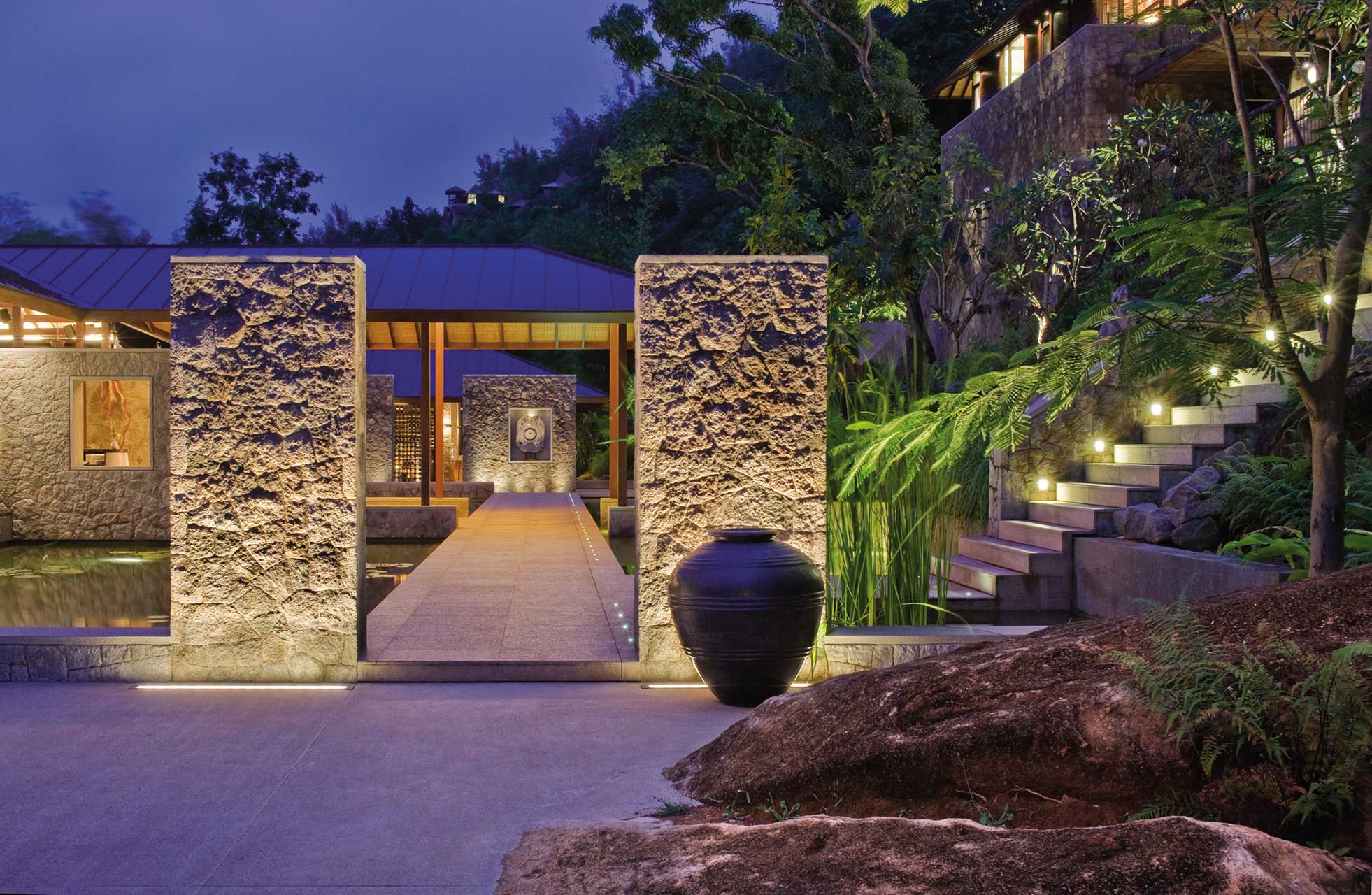 Modern stone hotel entrance lit at dusk, with large pottery, surrounded by lush greenery and illuminated stairs, creating a serene, inviting atmosphere.