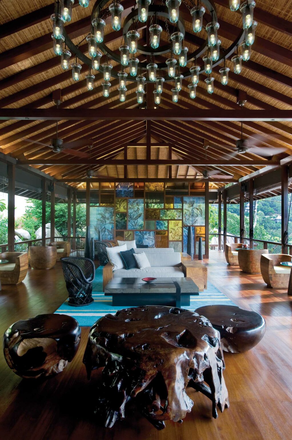 Spacious open-air hotel lounge with rustic wooden ceiling, large chandelier, and vibrant stained glass wall. Cozy sofa and unique sculpted wood tables. Relaxing, artistic atmosphere.