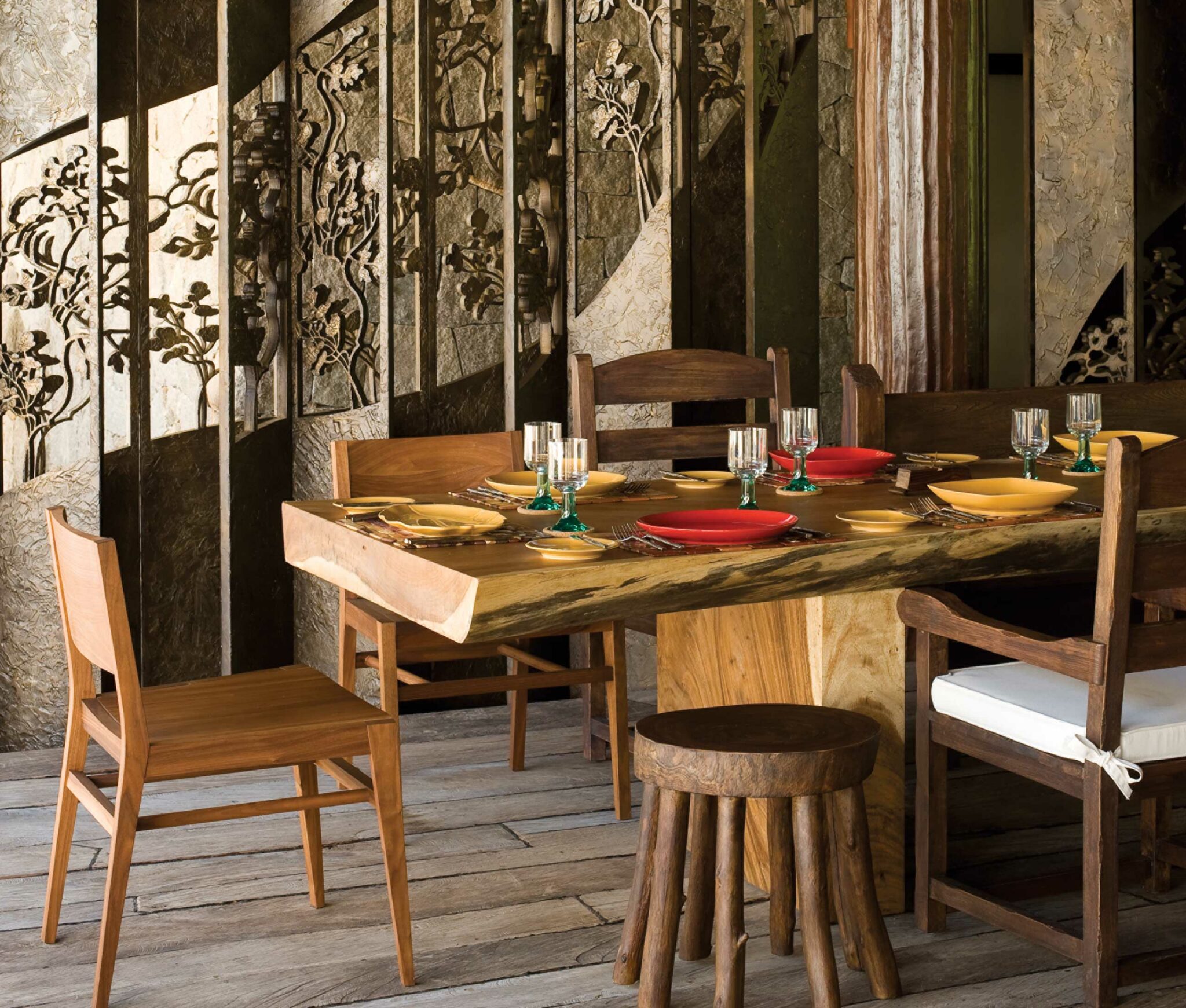 Rustic hotel dining area with a wooden table set with colorful red and yellow plates and clear glasses. Decorative wall panels feature intricate carvings. Warm and inviting atmosphere.