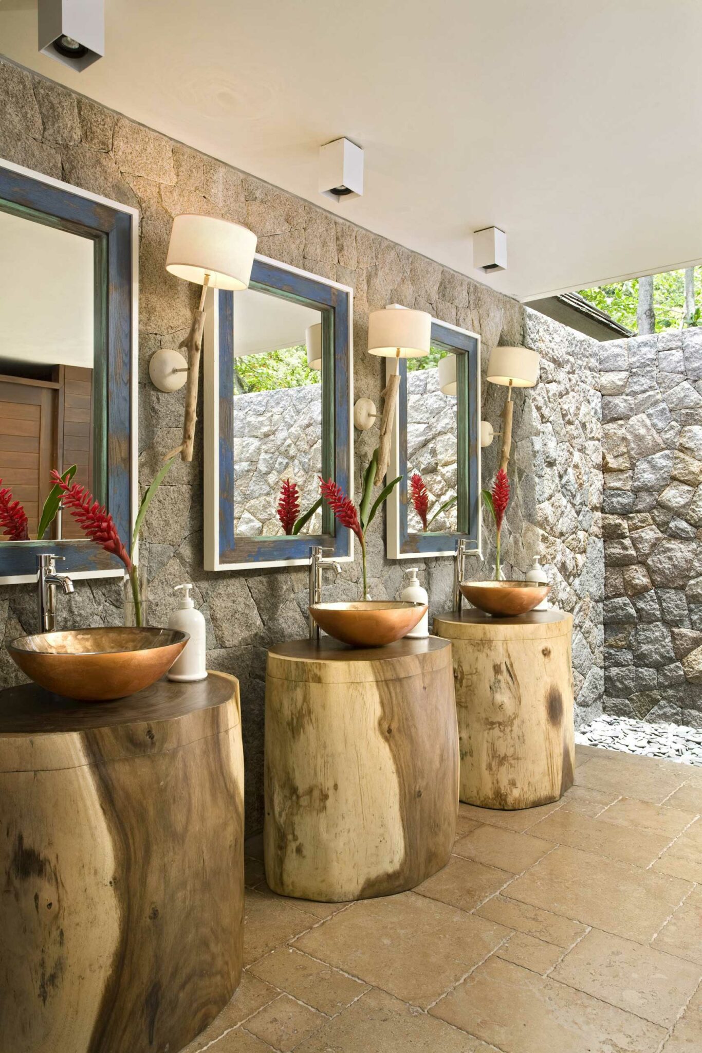 Rustic hotel bathroom with stone walls, featuring three wooden stump vanities with copper sinks. Each vanity has a red flower and a mirror above, exuding a natural and cozy feel.