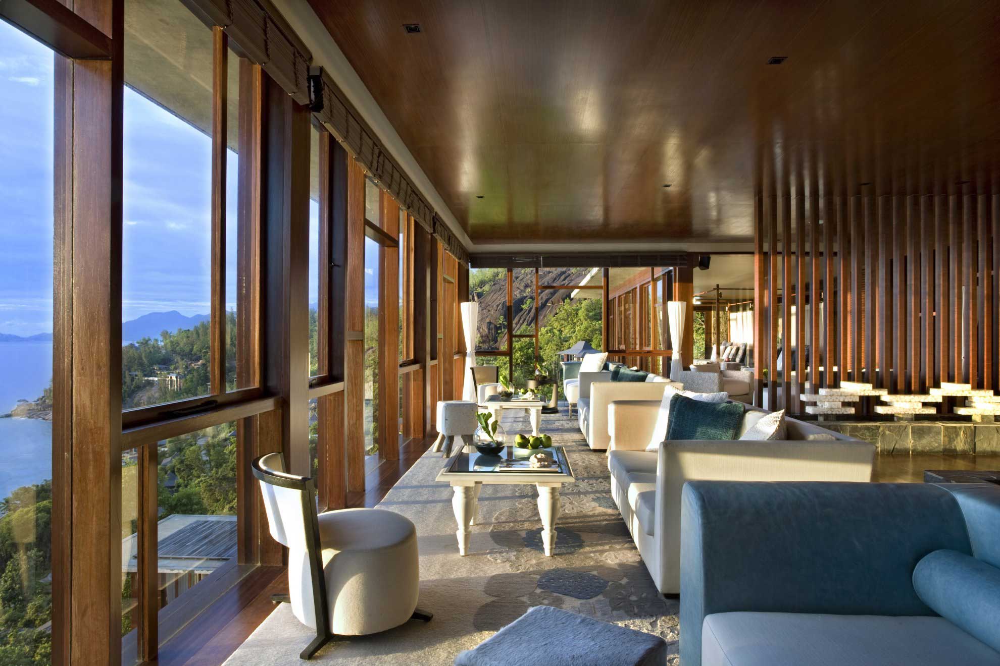 Luxurious open-plan hotel lounge with large windows offering a scenic view of mountains and sea. Soft white and blue-toned furnishings create a serene atmosphere.
