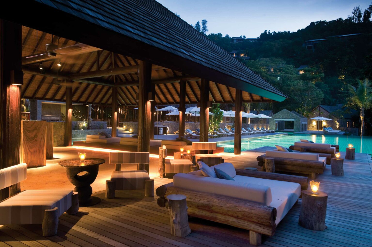 Luxurious open-air hotel lounge by a pool at dusk. Cozy sofas and wooden tables under a thatched roof, with warm lighting creating a serene, inviting atmosphere.