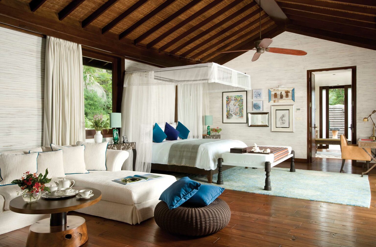 Luxurious hotel bedroom with a canopy bed draped in white, accented by teal and blue pillows. It features elegant decor, a cozy sofa, and natural light pouring in.